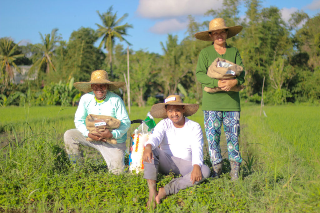 Three Filipino farmers on a sunny, green field, posing with some of their rice produce.