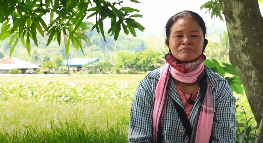 An elderly female farmer sitting under the shade of the tree, with a green field in the background.